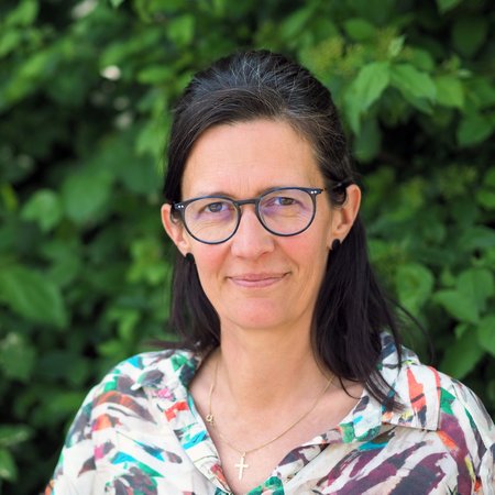 Eine Frau mit langen dunklen Haaren und einer Brille vor Blattwerk