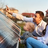 Zwei Personen sitzen vor einer Solarzelle und zeigen auf etwas, das sich ein Kind ansehen soll