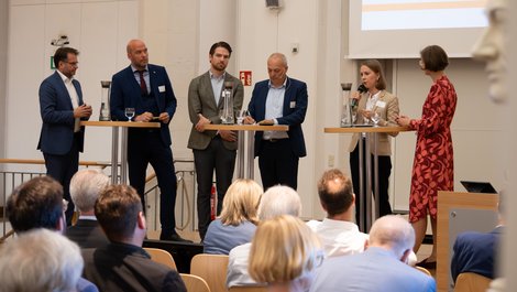 Göttinger Energietagung: Vier Männer und zwei Frauen stehen an Stehtischen und diskutieren miteinander. Vor ihnen sitzen Menschen auf Stühlen und hören zu.